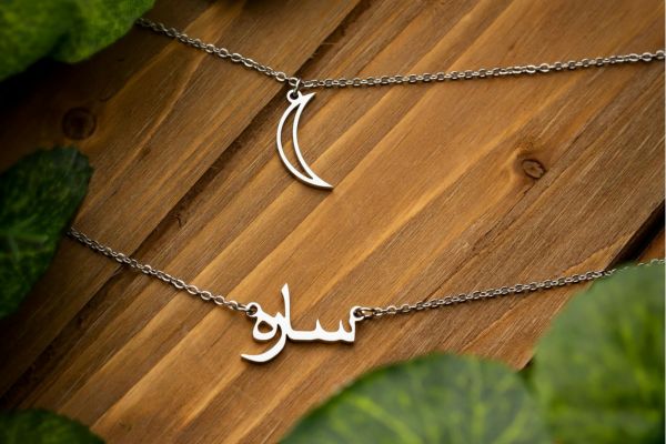 Two silver necklaces on wood, one with a crescent moon, the other with Arabic script.
