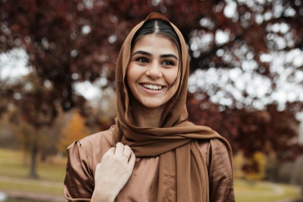 Smiling person wearing a brown headscarf, standing outdoors with trees in the background.