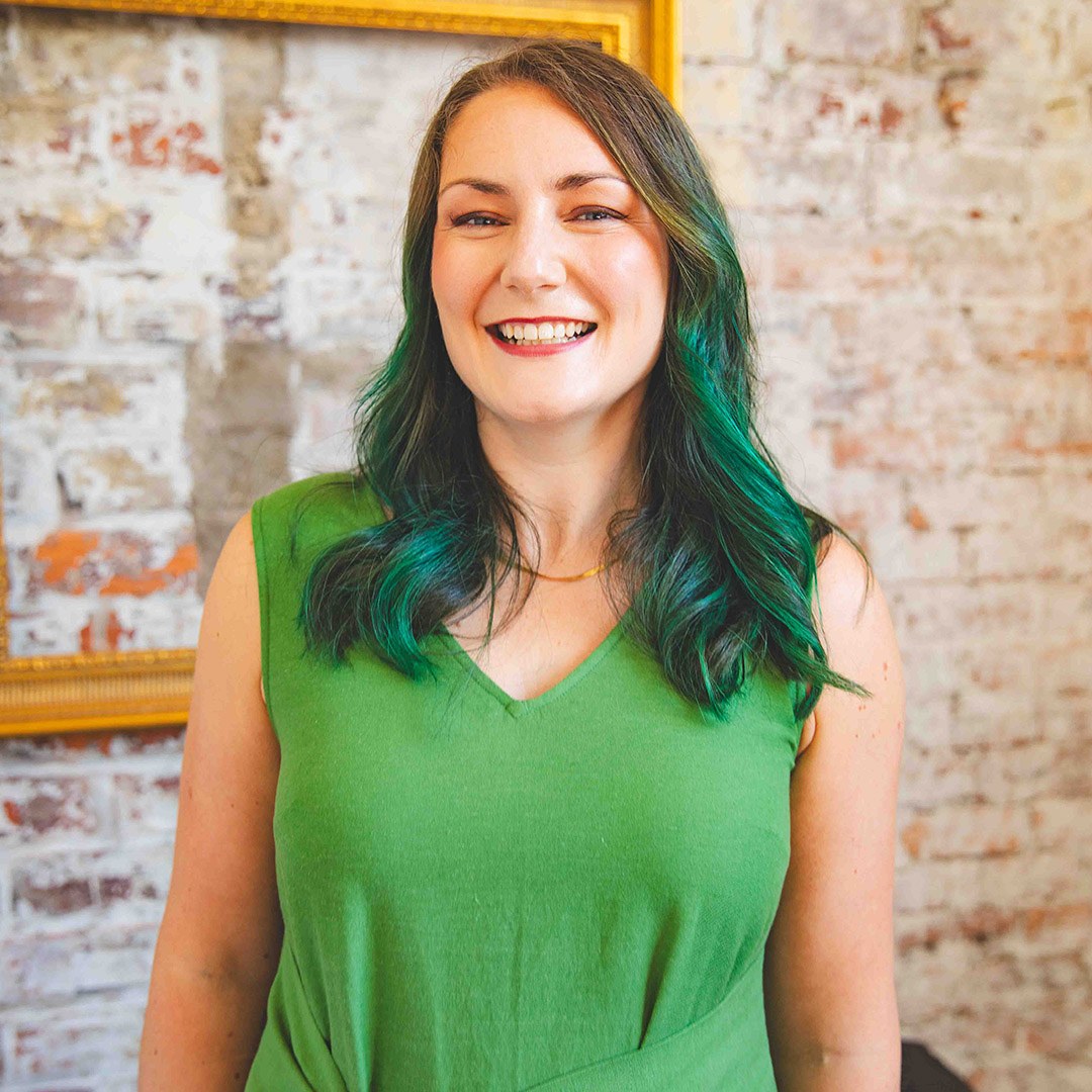 A smiling woman with green hair and a green top stands in front of a brick wall.