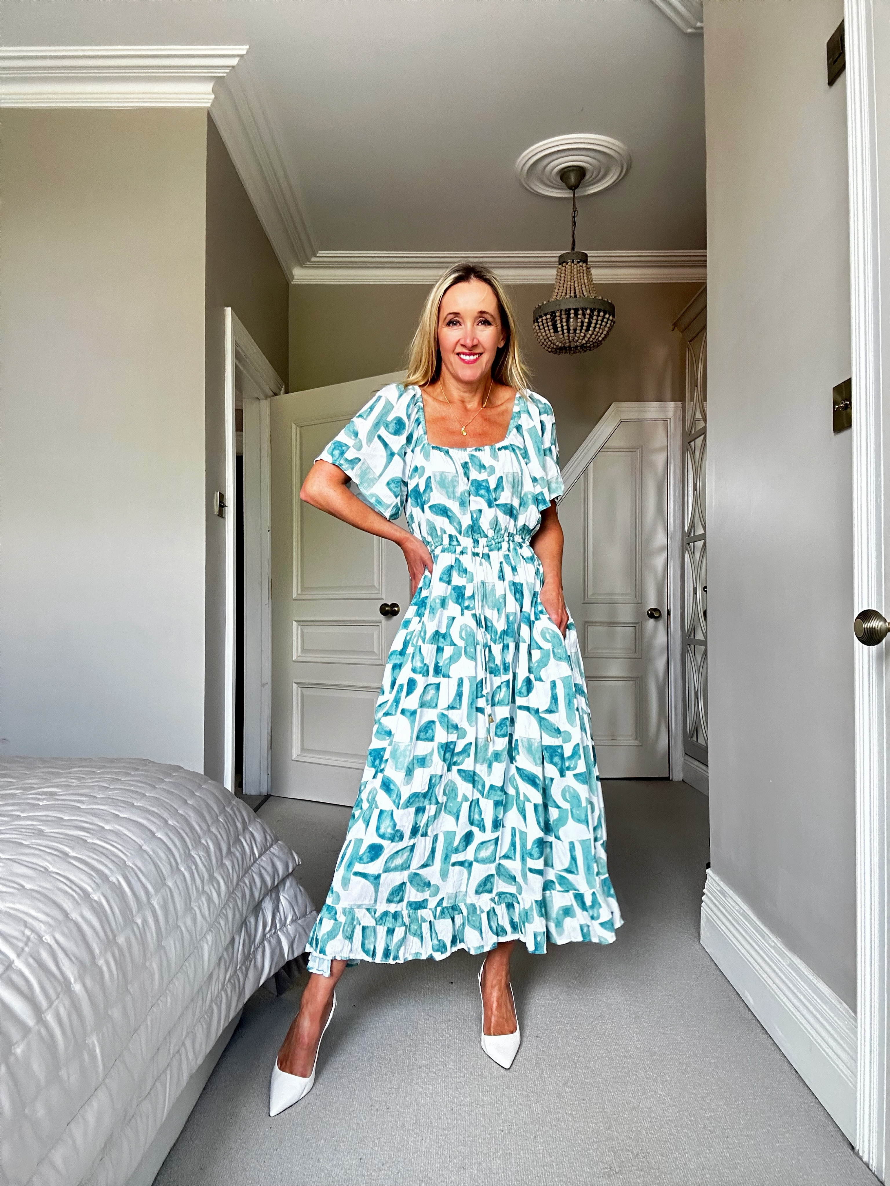 Woman in a patterned dress standing in a bedroom with a chandelier.