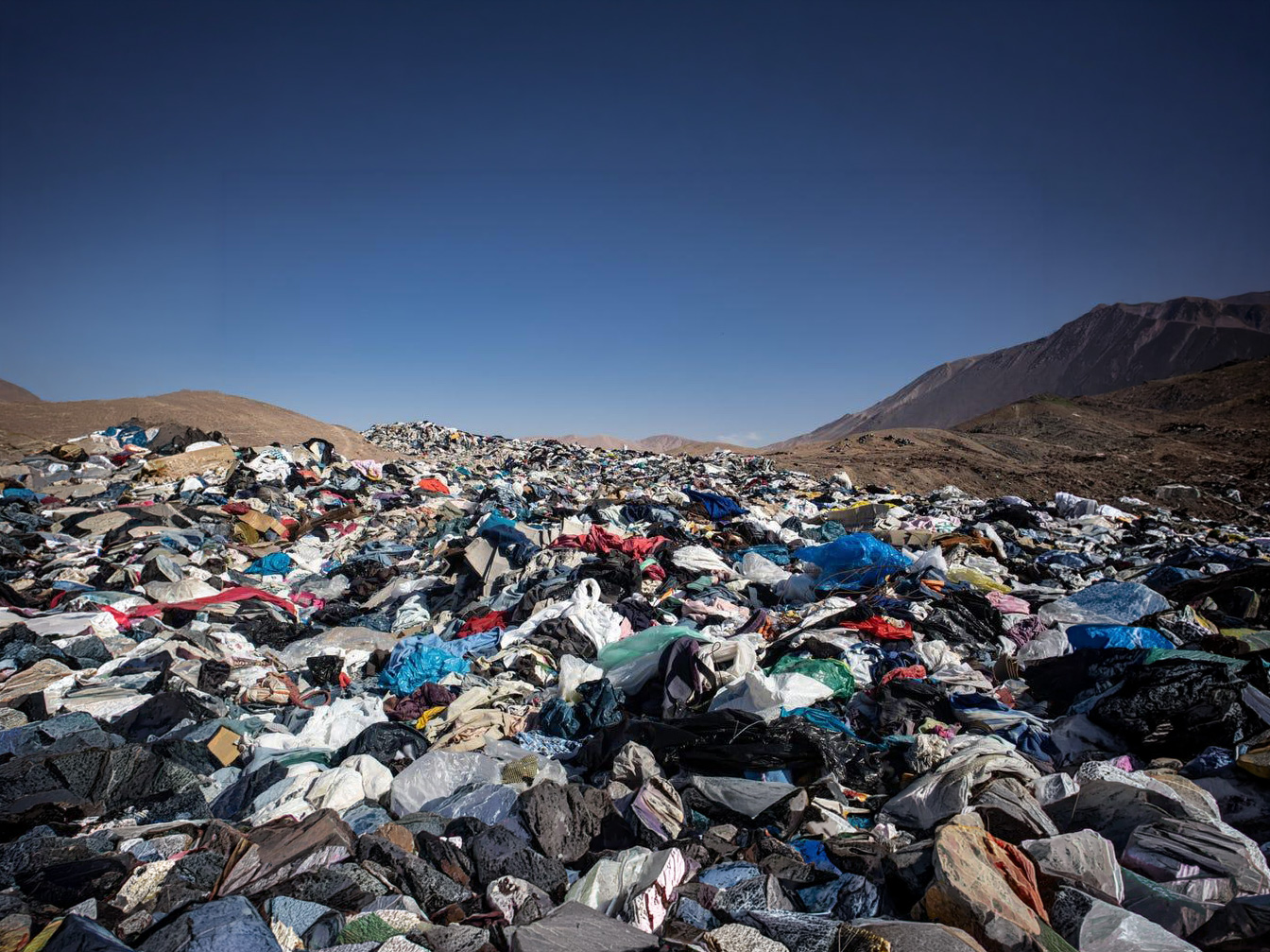 A vast pile of discarded clothing fills a desert landscape, with mountains in the background.