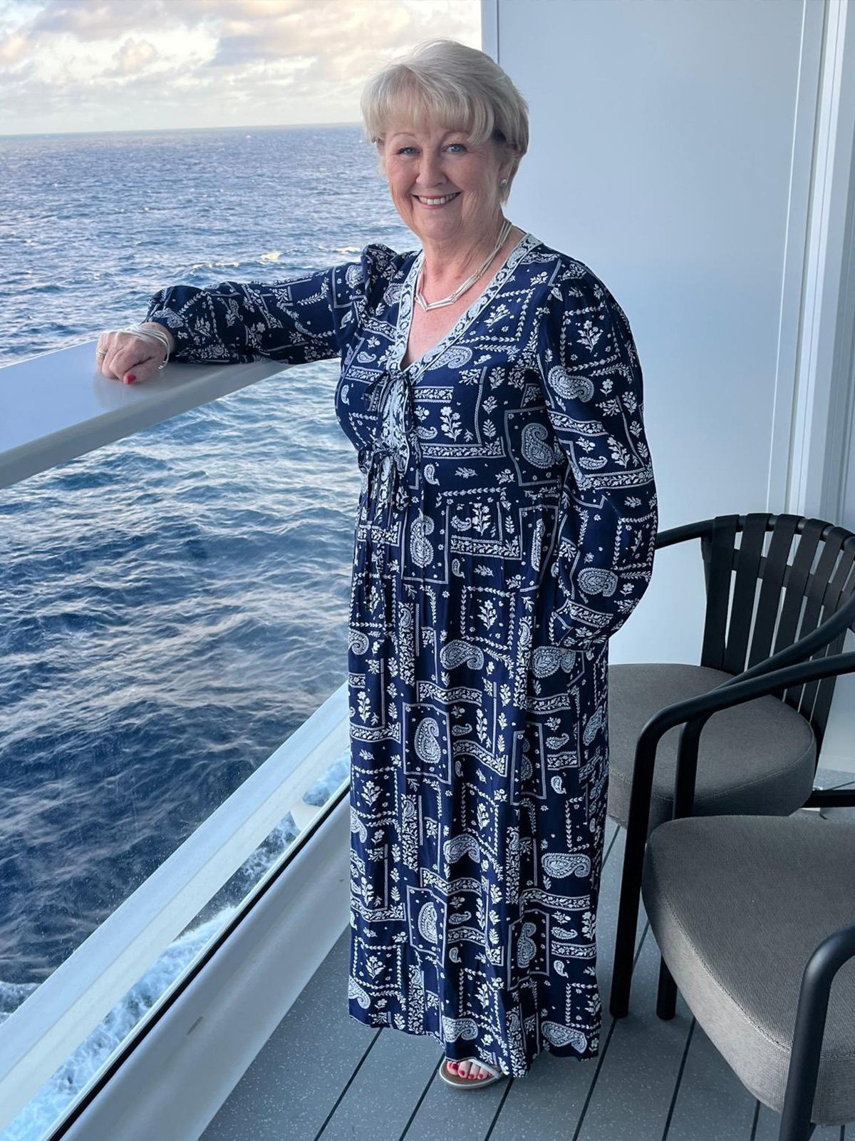 A woman in a long blue and white patterned dress smiles on a balcony overlooking the ocean.