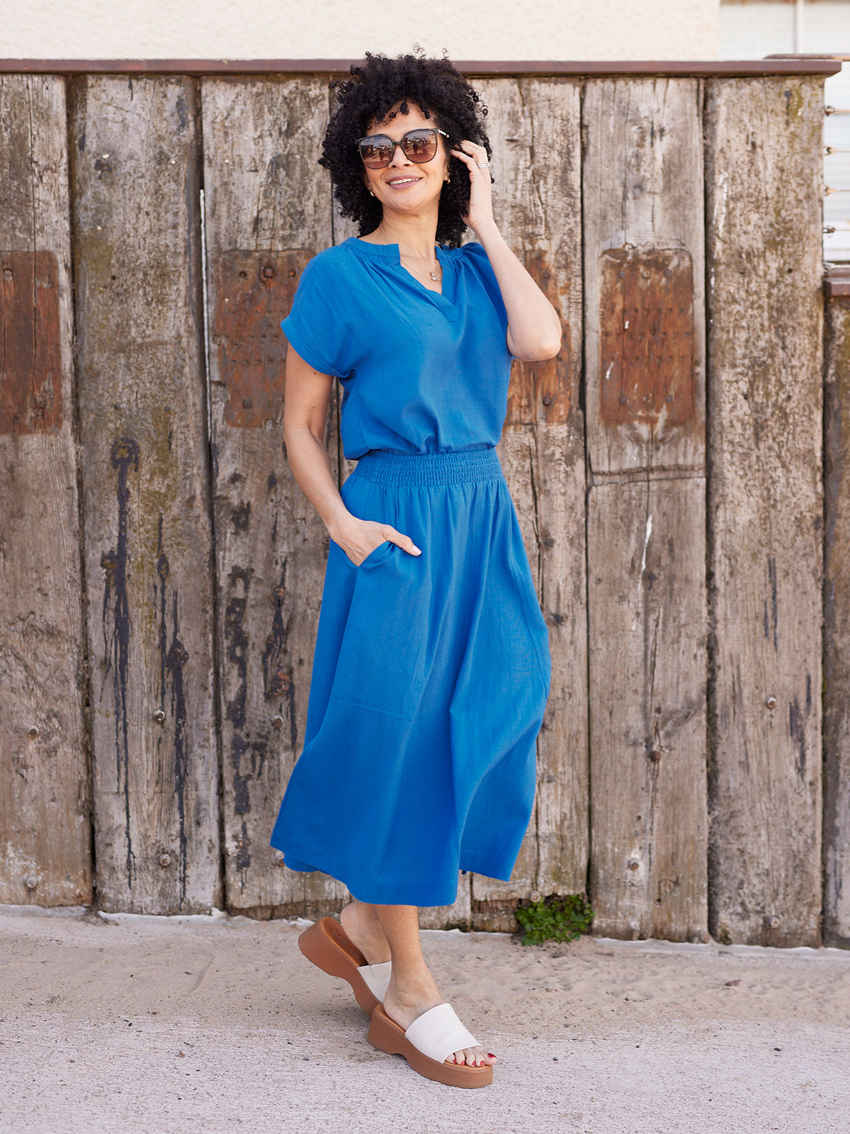 Person in blue dress and sunglasses standing by a wooden fence.