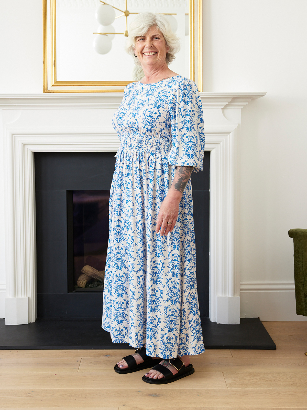 Person in a blue floral dress standing by a fireplace, smiling.
