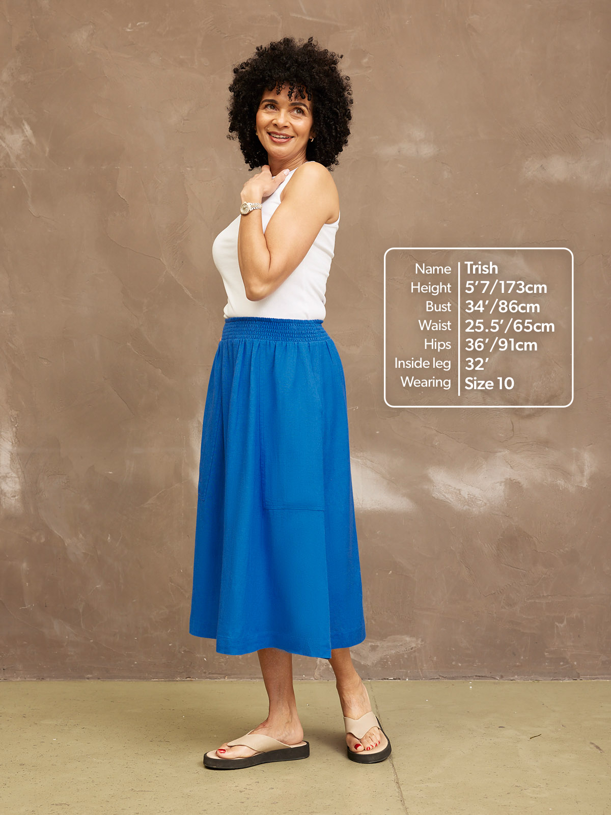 Woman in white top and blue skirt standing against a textured wall.