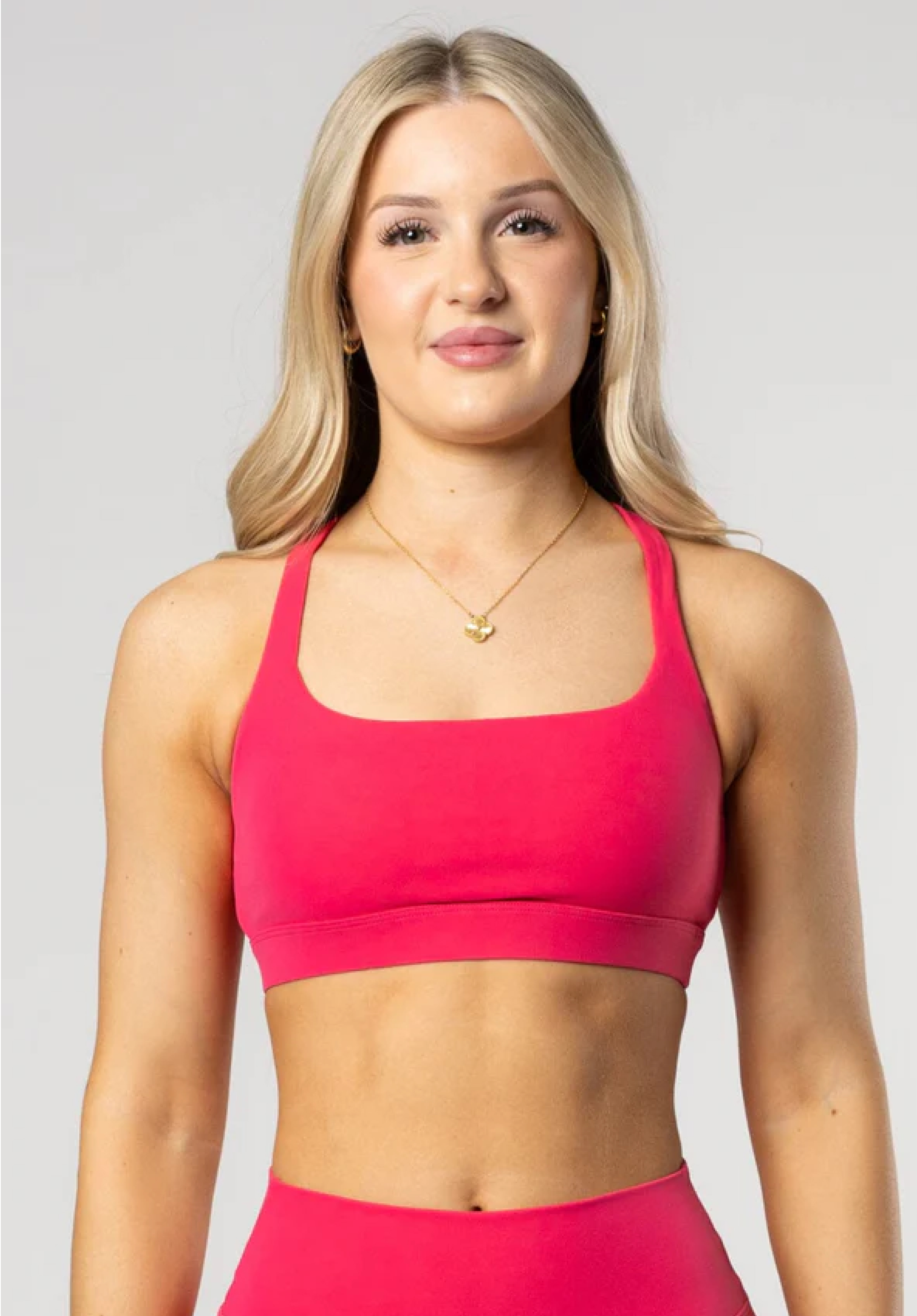A woman with blonde hair models a pink sports bra and a gold necklace against a grey background.