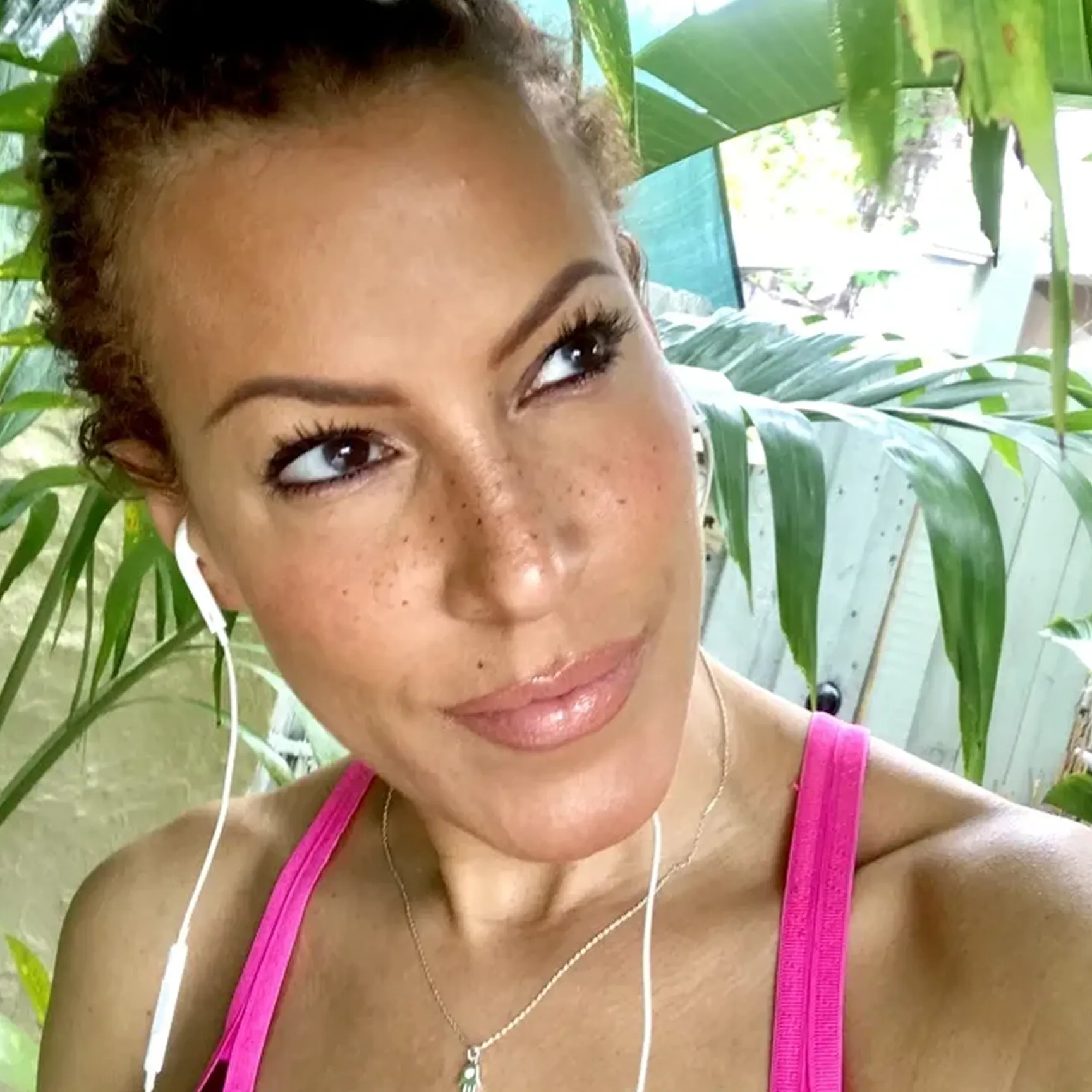 A woman with freckles wearing a pink tank top and earbuds looks up and to the side.