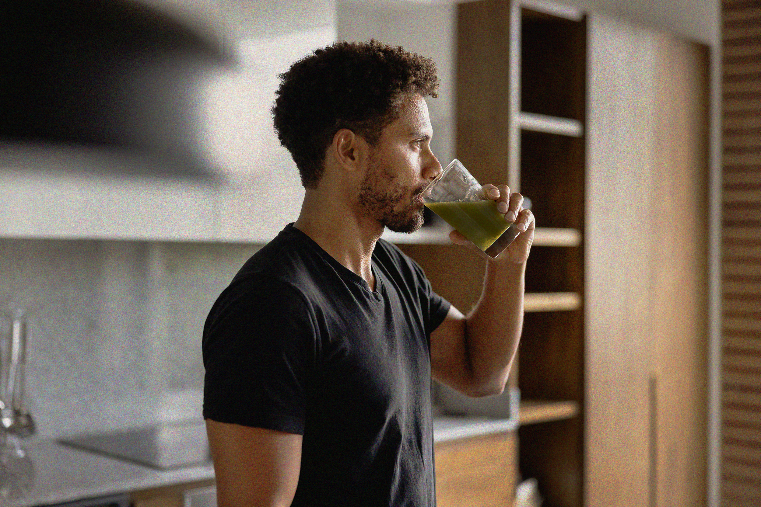 A man in a black t-shirt stands in a kitchen and drinks a green beverage from a glass.