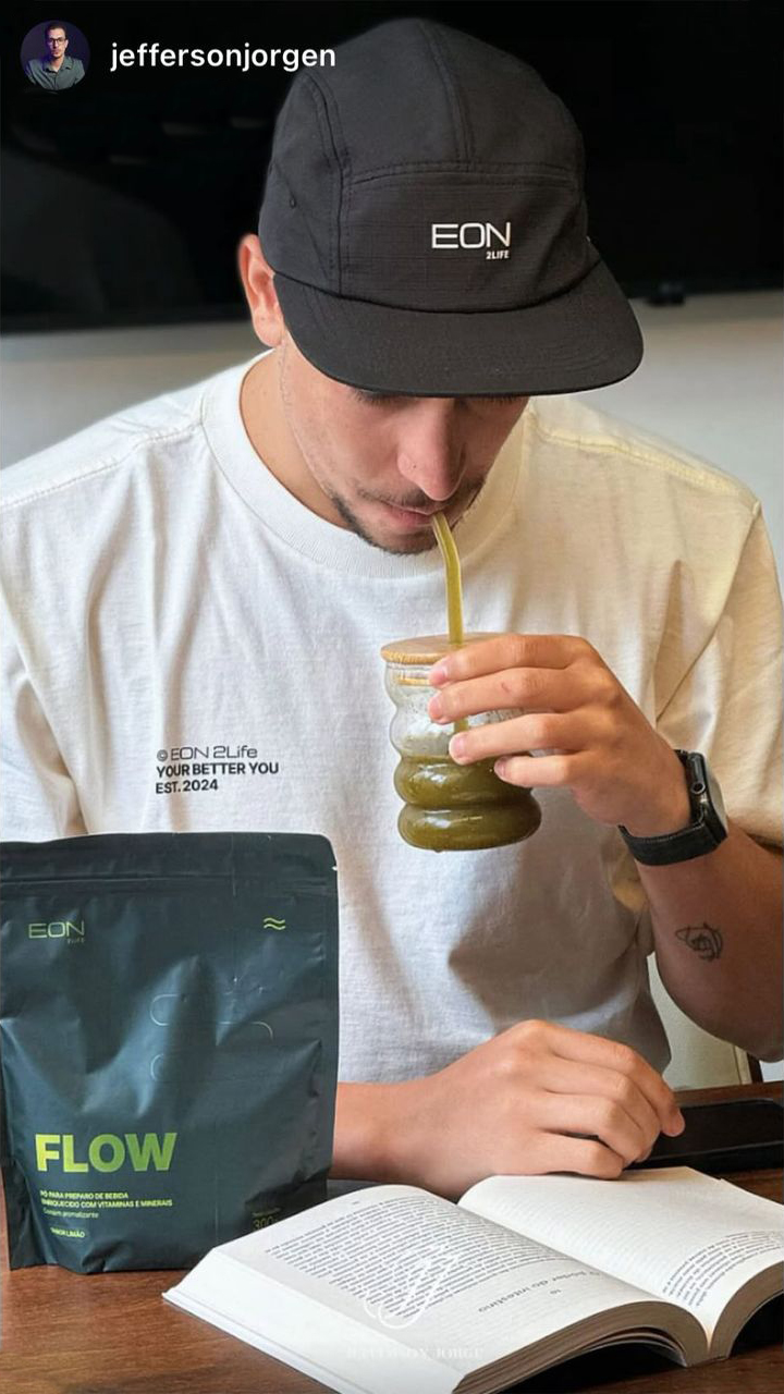 A man in a cap and t-shirt drinks a green beverage through a straw while reading a book.