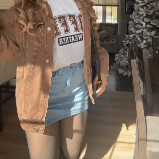 Person in denim skirt and brown jacket inside a sunlit room.