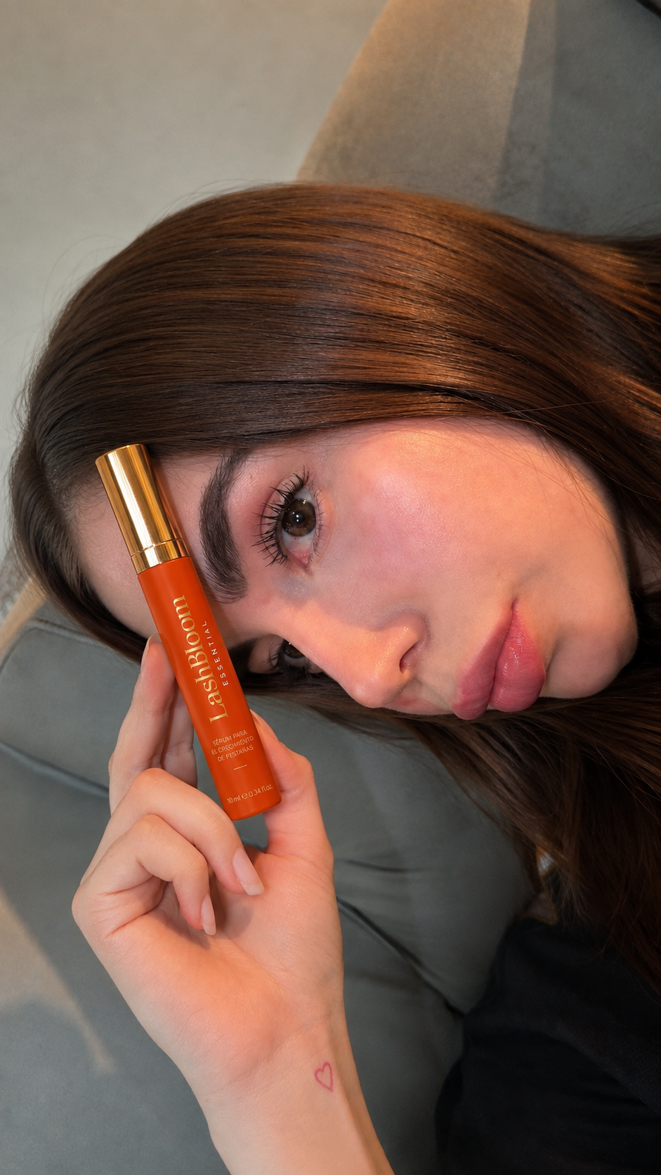 A woman with long brown hair holds an orange tube of LashBloom eyelash serum near her face.