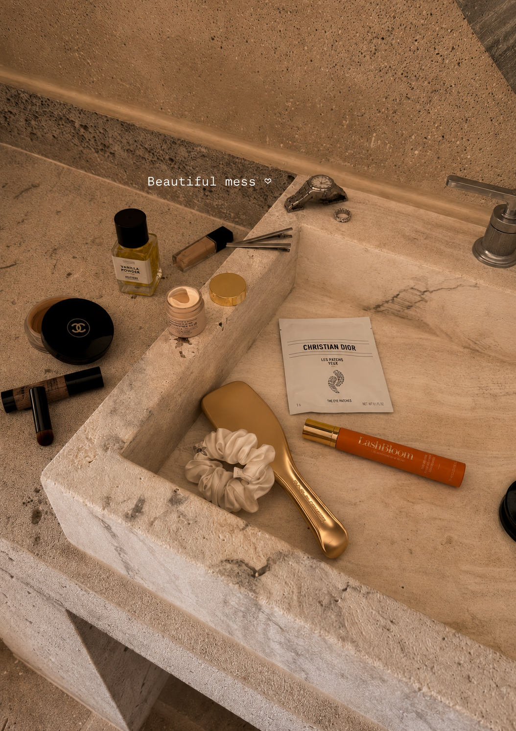 Various makeup and beauty products, a watch, and a hairbrush scattered on a stone sink.