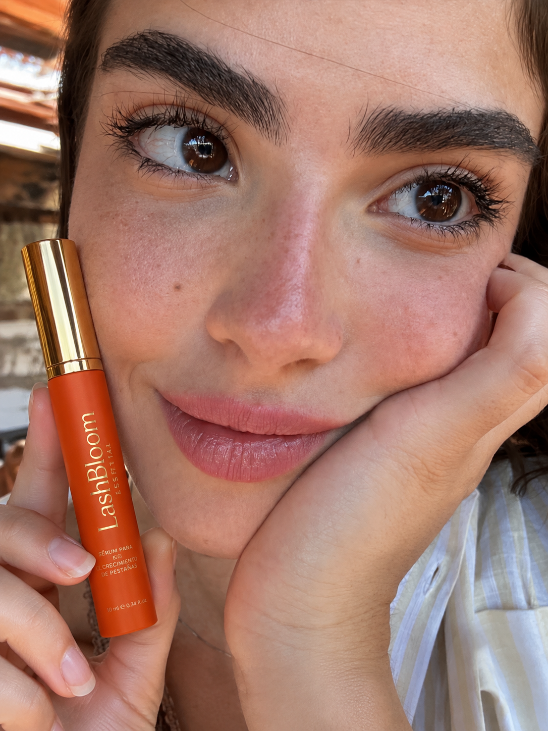 A close-up of a woman holding a small orange tube of LashBloom eyelash serum next to her face.