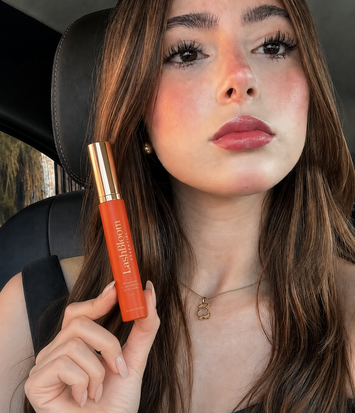 A woman with long brown hair sits in a car, holding up an orange tube of LashBloom Essential serum.
