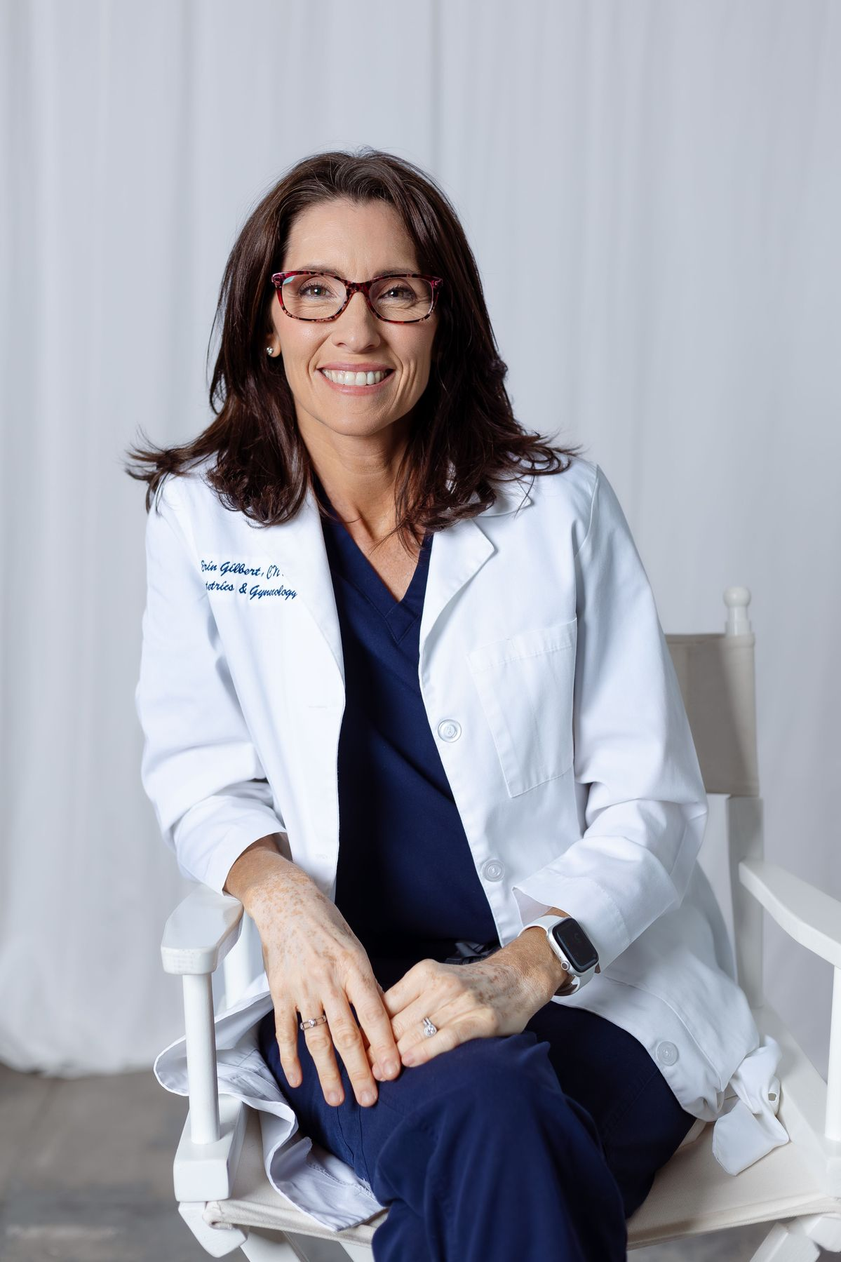 A smiling woman with glasses, wearing a lab coat over scrubs, sits in a white director's chair.
