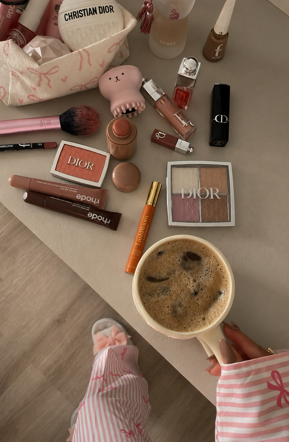 An overhead view of various makeup products on a table next to a person holding a mug of coffee.