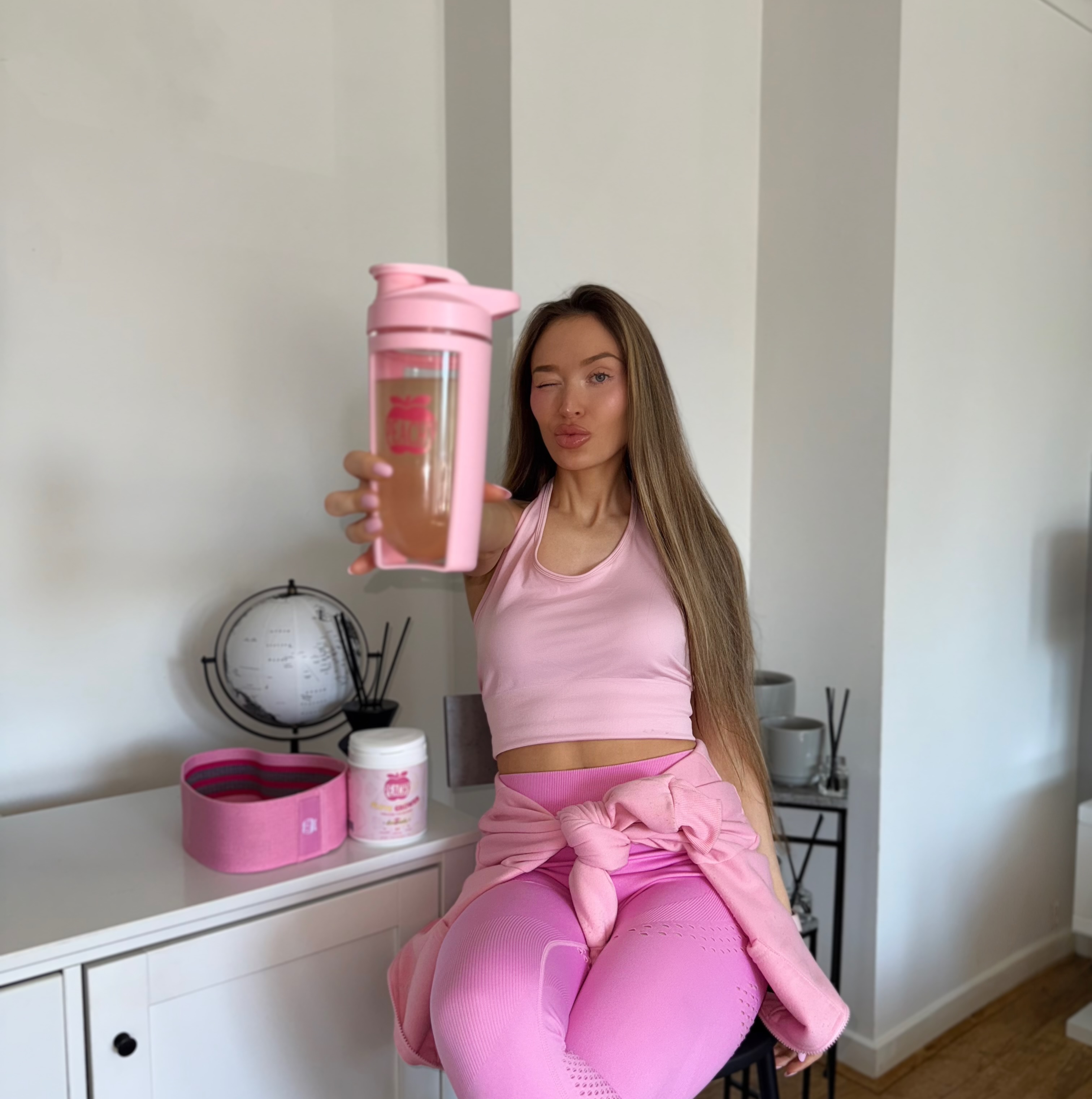Person in pink workout attire holding a pink shaker bottle, sitting on a white cabinet.