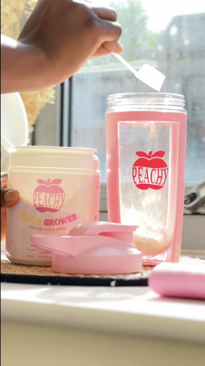 A person prepares a pink drink using a pink container labeled 'Peachy Glute Grower' on a windowsill.
