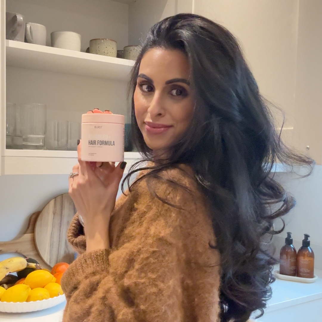 A woman with long dark hair holds a pink container of Burst Hair Formula in a kitchen.