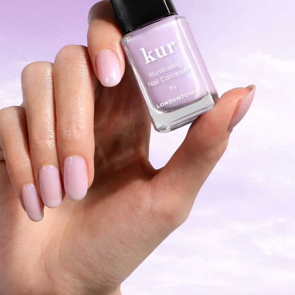 A hand with a sheer lavender manicure holds a bottle of kur Illuminating Nail Concealer.