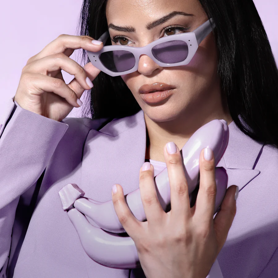 A woman in a lavender suit and sunglasses holds a bunch of lavender-colored bananas against a lavender background.