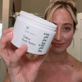A woman holds up a white jar of Nimi Body Tallow Body Butter and smiles at the camera.