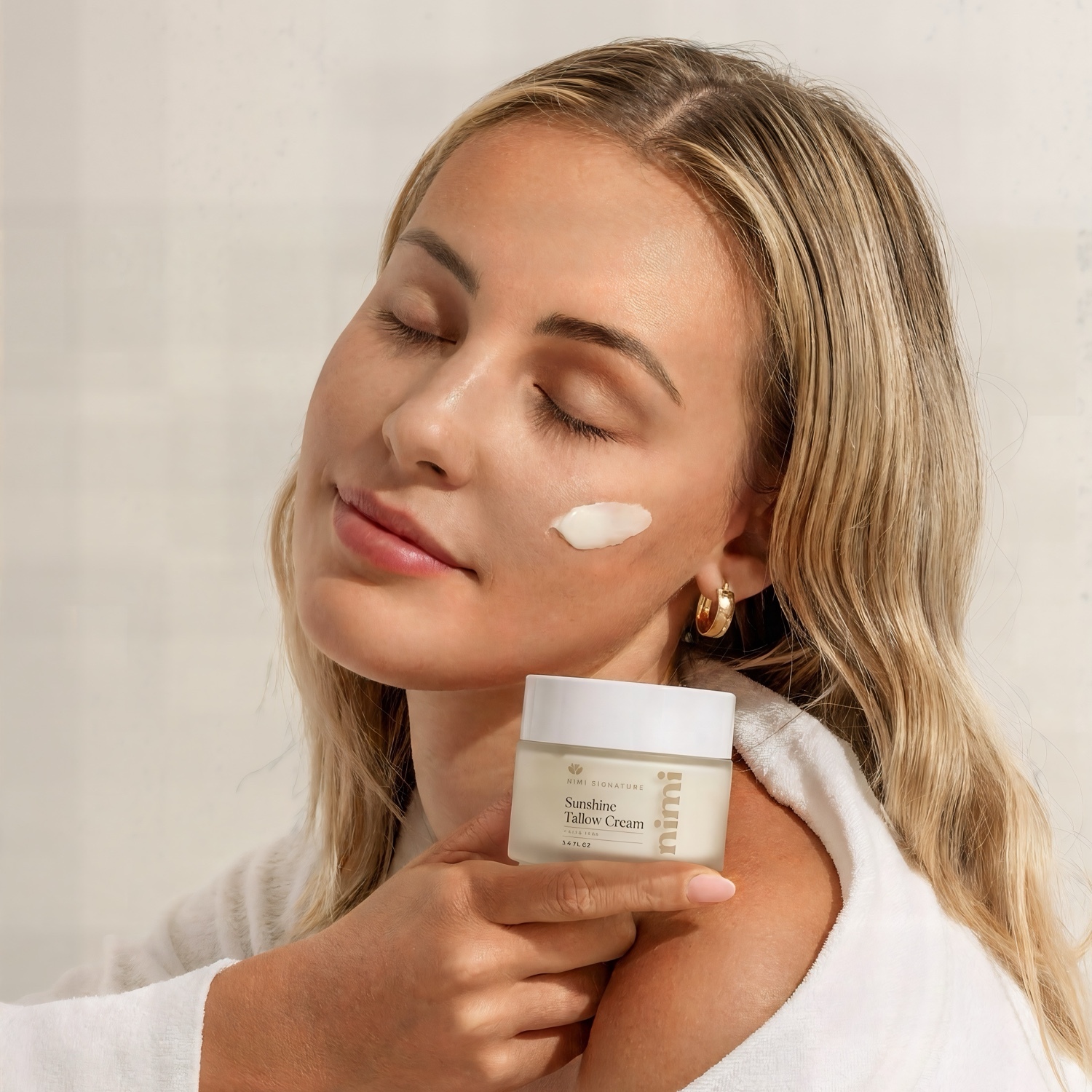 A woman in a white bathrobe holds a jar of cream with a dab of it on her cheek.