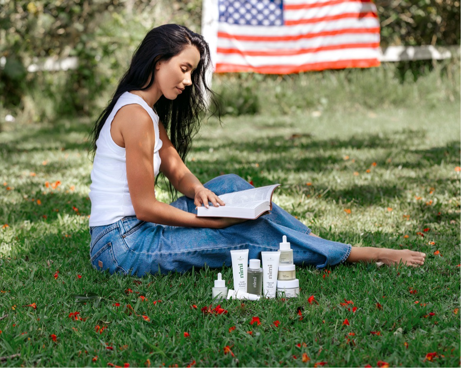 A woman sits barefoot on grass reading a book, with beauty products and an American flag in the background.