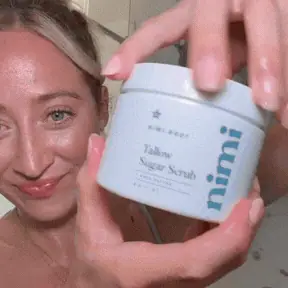 A smiling woman holds a white jar of Nimi Tallow Sugar Scrub.