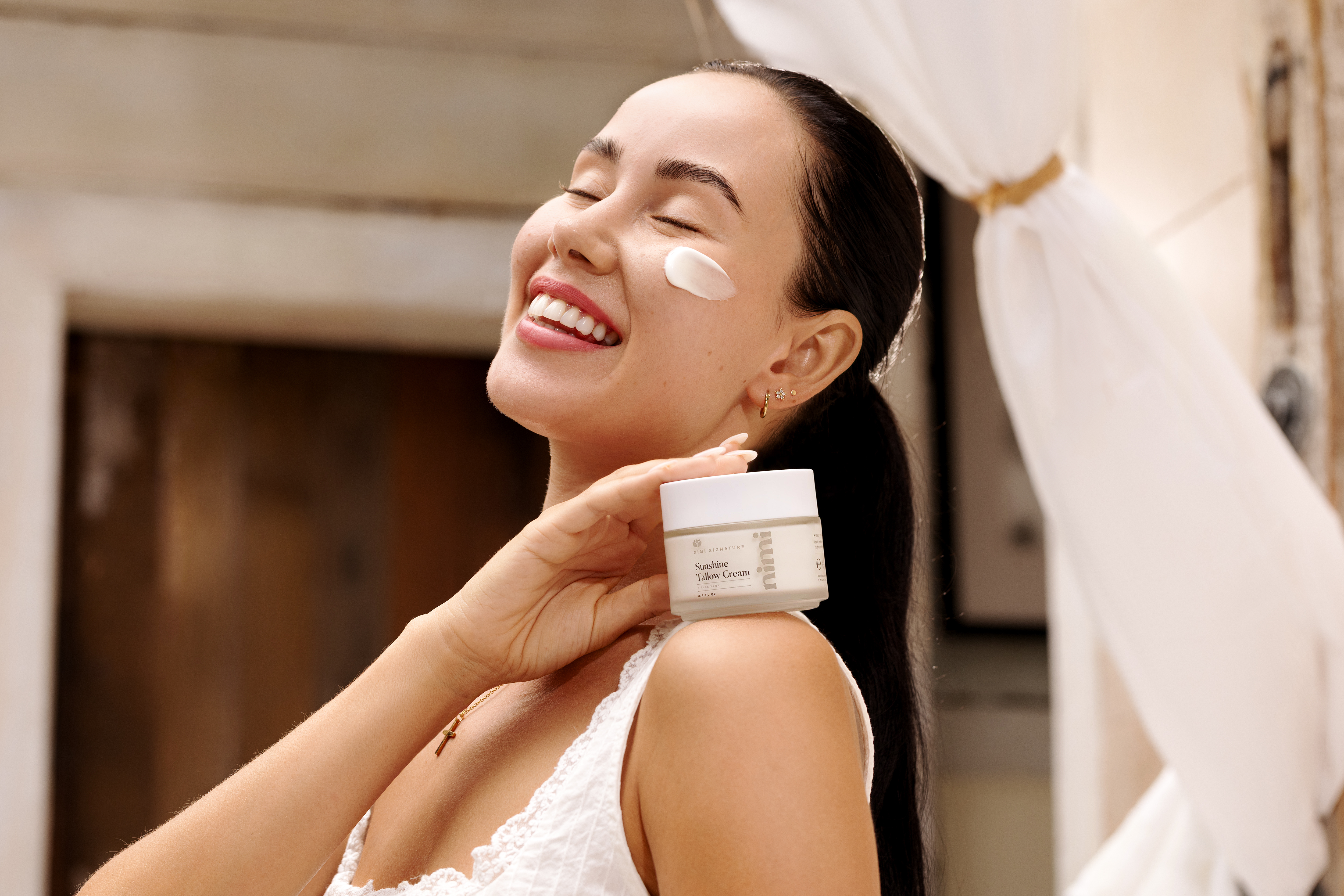 A smiling woman with eyes closed has a dab of face cream on her cheek while holding the jar.