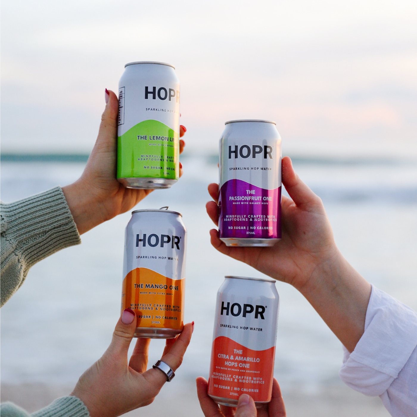 Four hands holding different flavored HOPR sparkling hop water cans.