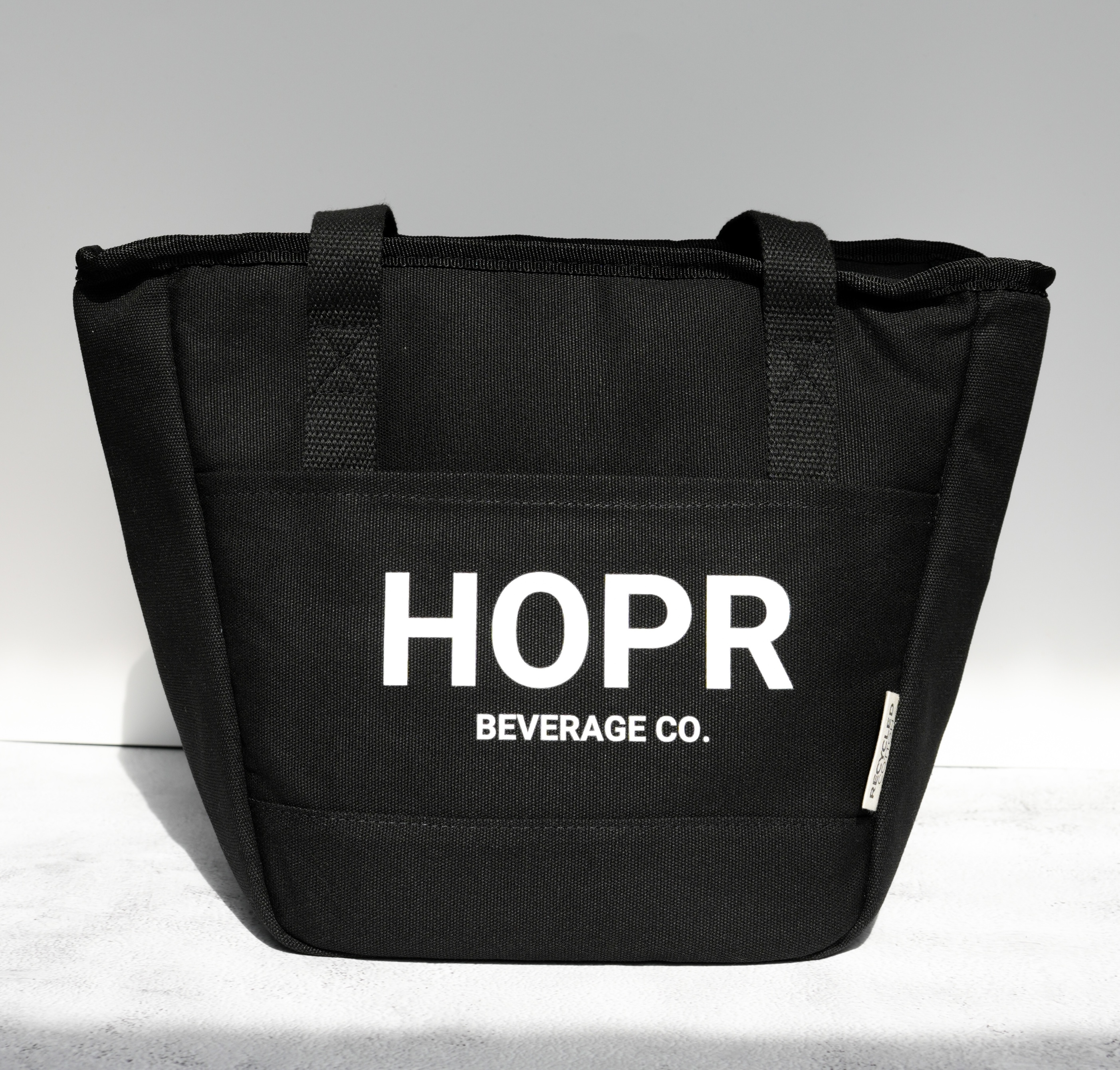 A black tote bag with 'HOPR BEVERAGE CO.' printed in white text on a light gray surface.