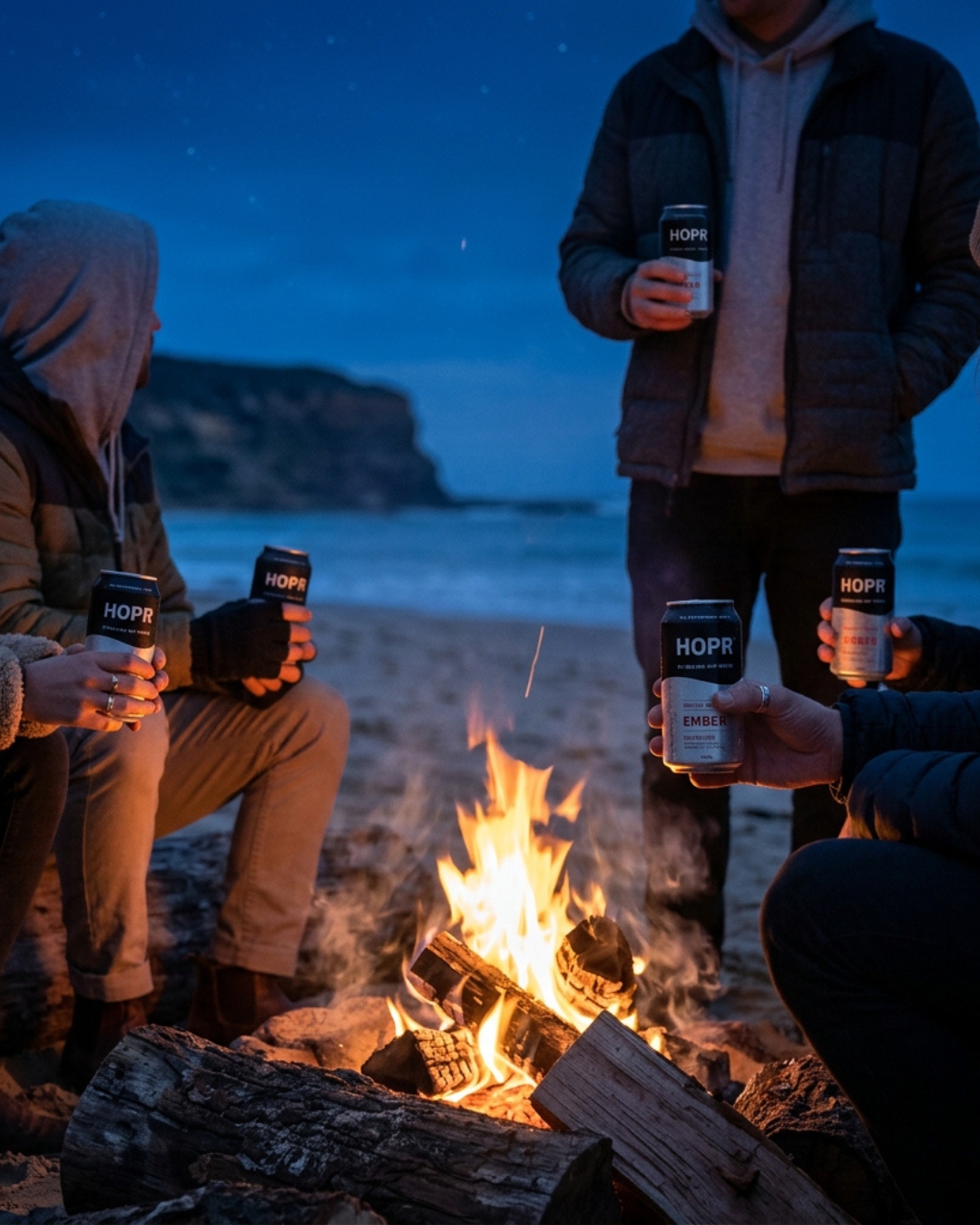 A group of people sit around a campfire on a beach at night, holding beverage cans.