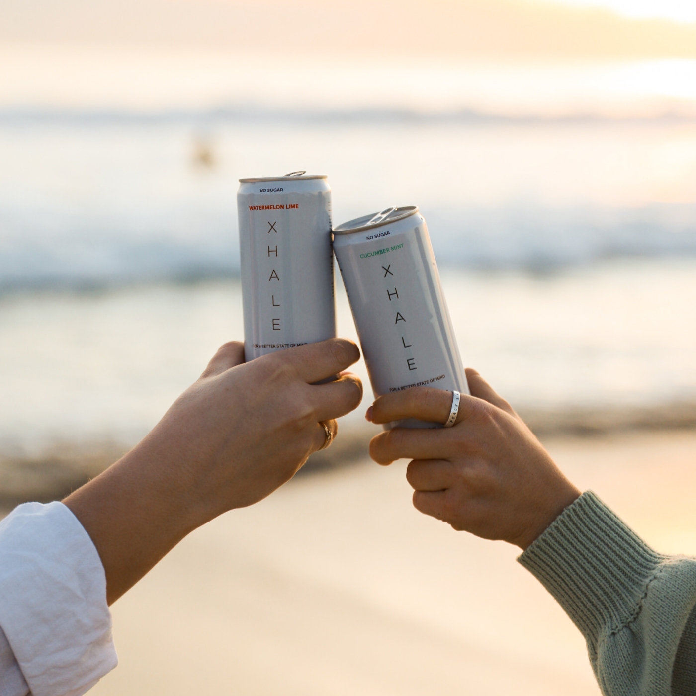 Two hands holding cans of XHALE beverage by the beach.