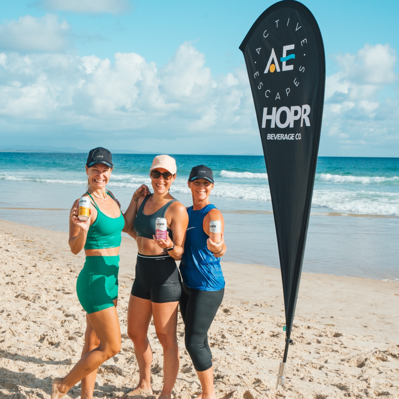Three women holding cans on a beach beside a flag with 'Active Escapes' logo.