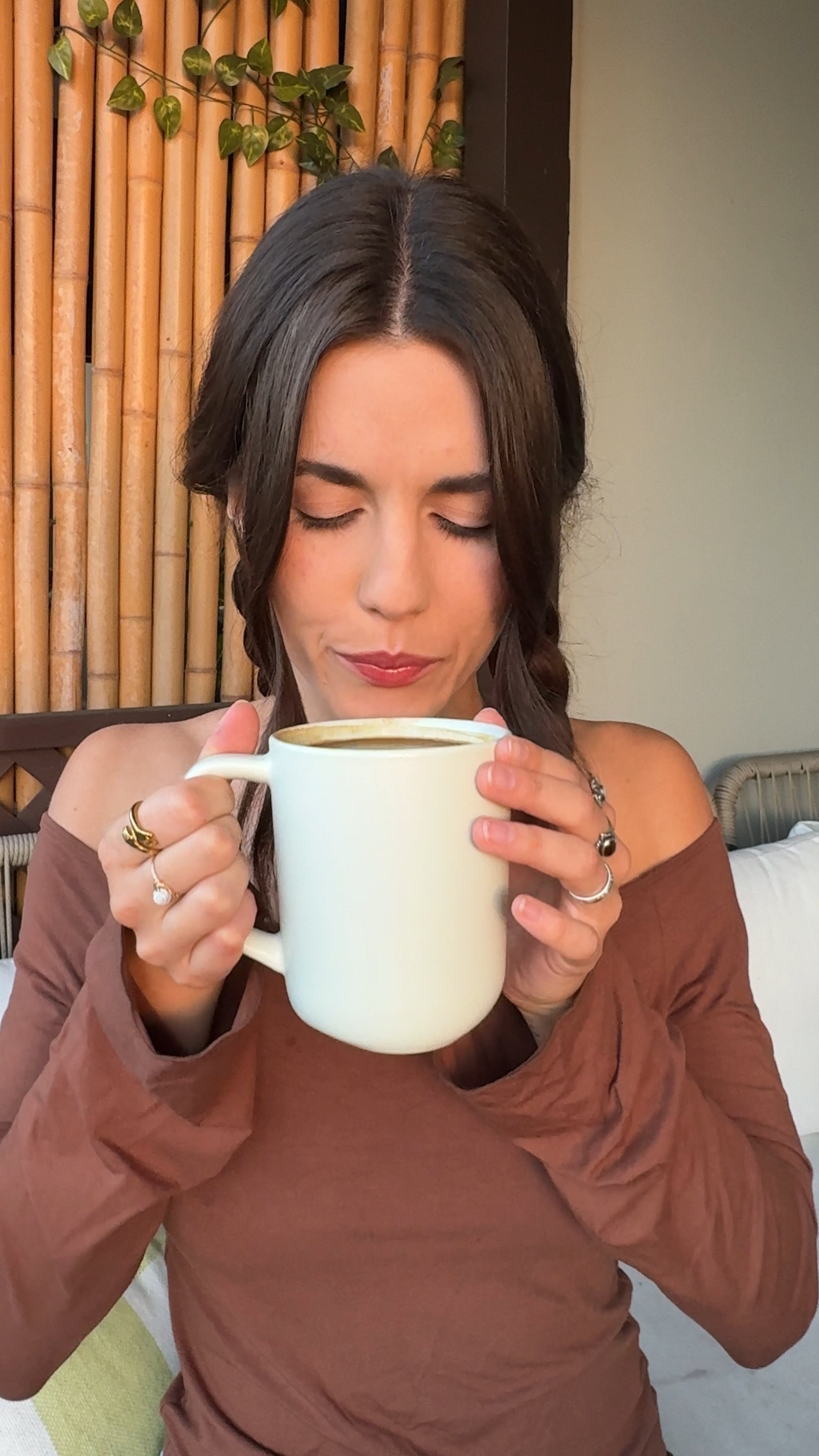 A woman with dark hair, eyes closed, smelling a drink from a large white mug.