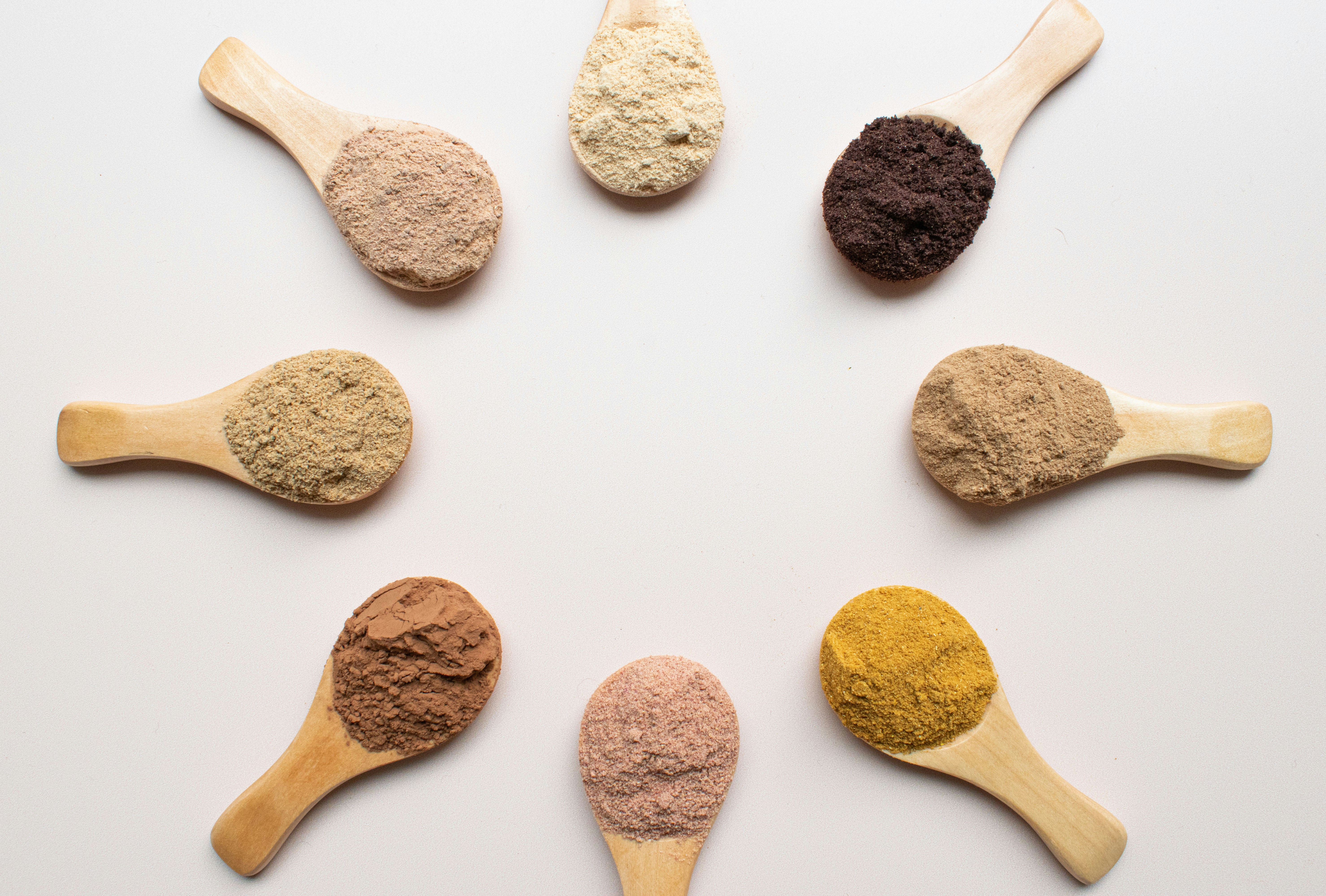 Wooden spoons with various colored powders arranged in a circle on a white background.