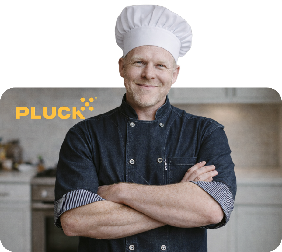 A smiling chef wearing a white hat and denim jacket stands with his arms crossed in a kitchen.