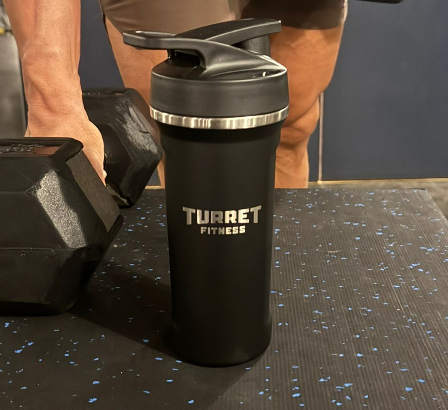 Black fitness bottle labeled 'TURRET FITNESS' beside a dumbbell in a gym setting.