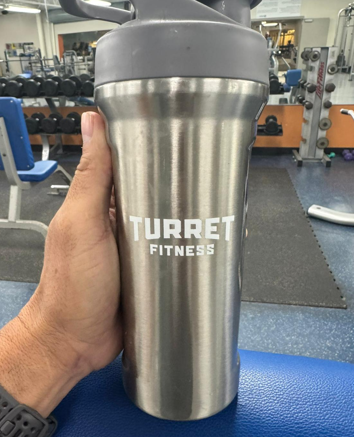 Stainless steel tumbler with 'Turret Fitness' logo in a gym setting.