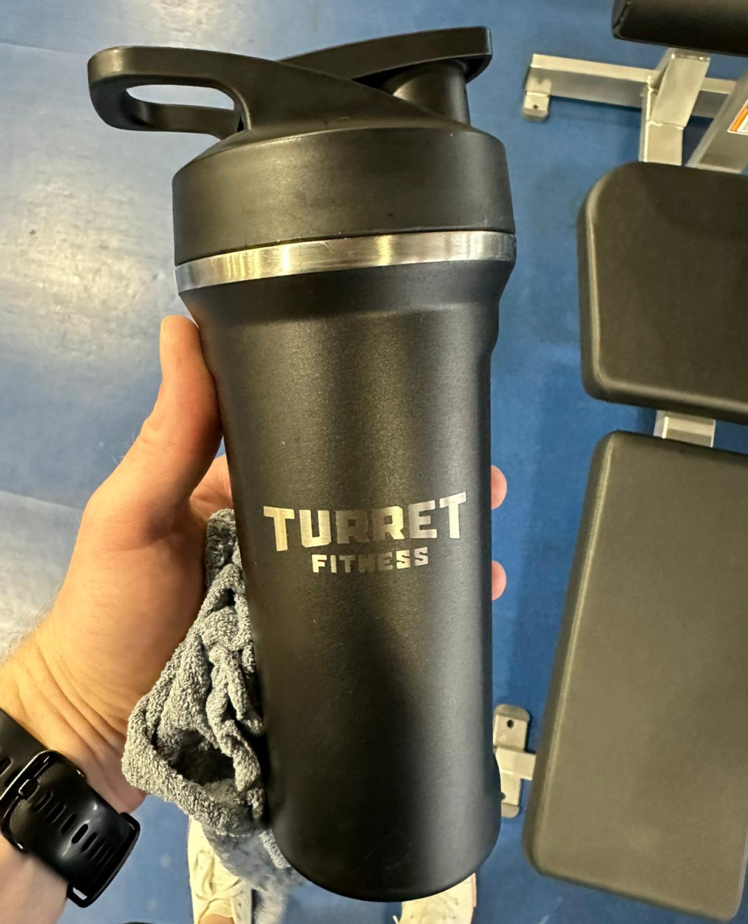 Hand holding a black shaker bottle labeled 'TURRET FITNESS' near gym equipment.