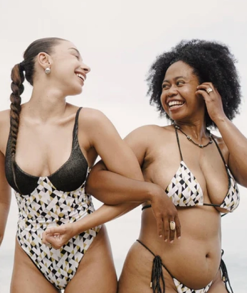 Two women in patterned swimsuits smiling and linking arms on a beach.