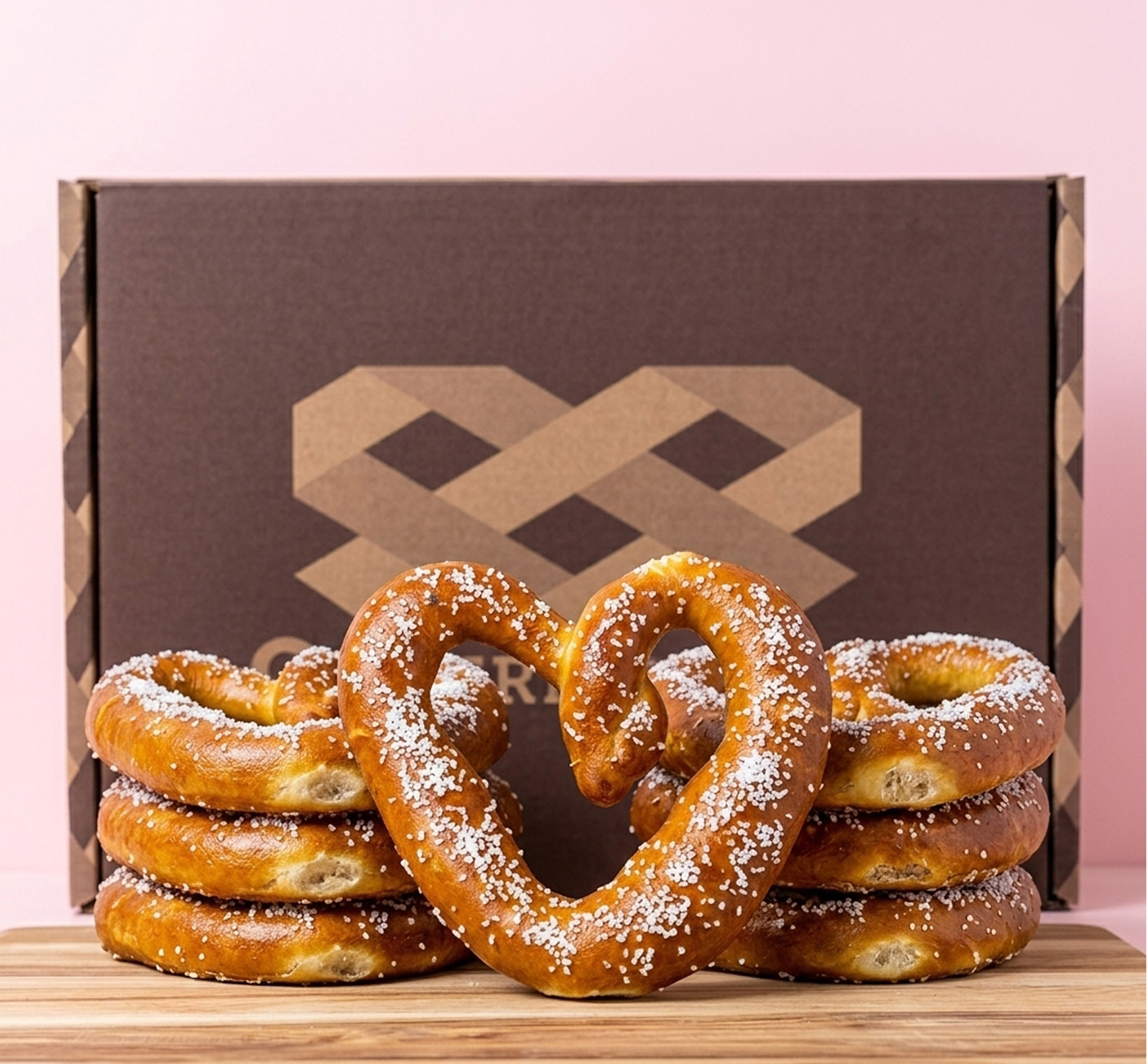 A heart-shaped soft pretzel and stacks of pretzels sit on a wooden board in front of a box.
