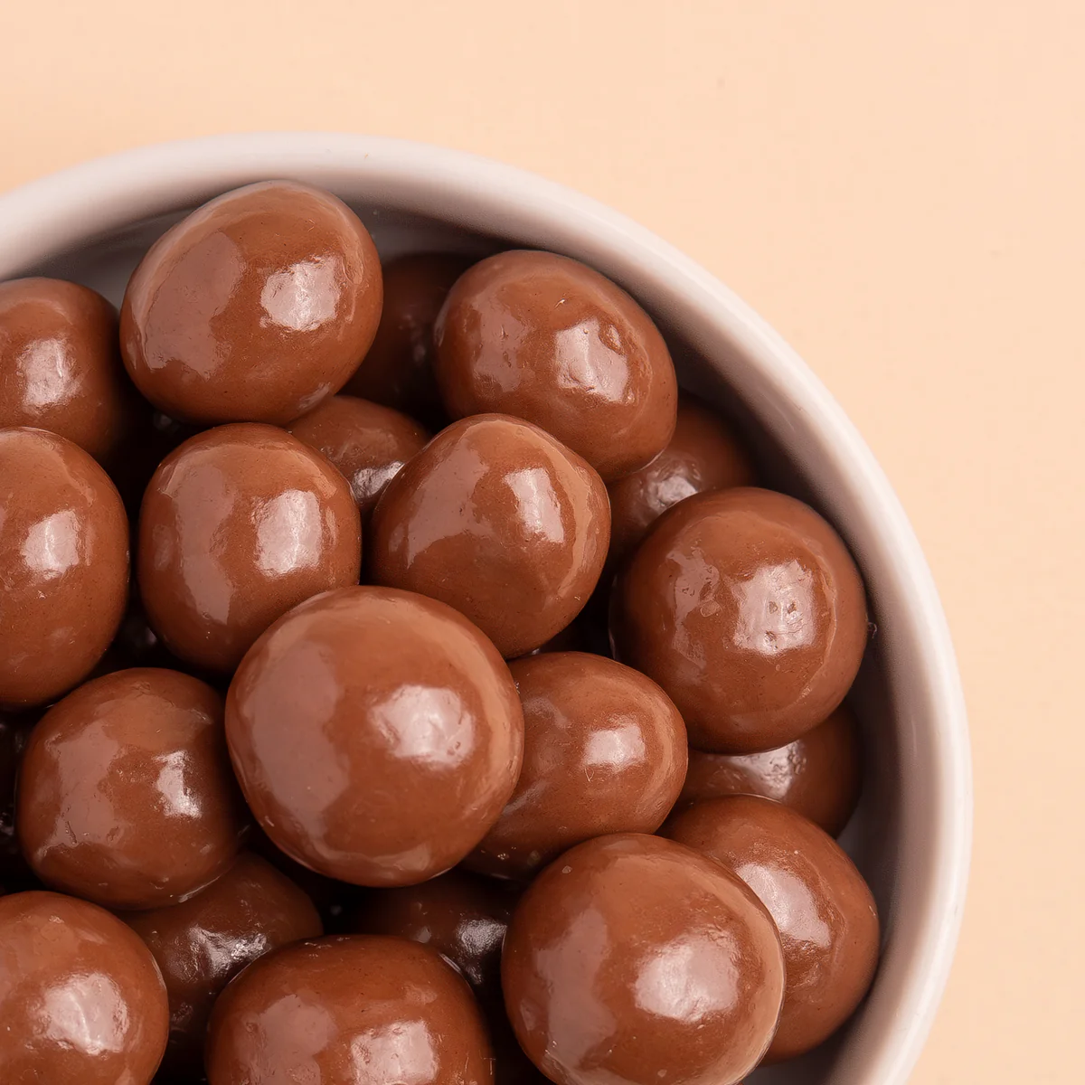 A close-up of a white bowl filled with shiny, round milk chocolate candies against a tan background.