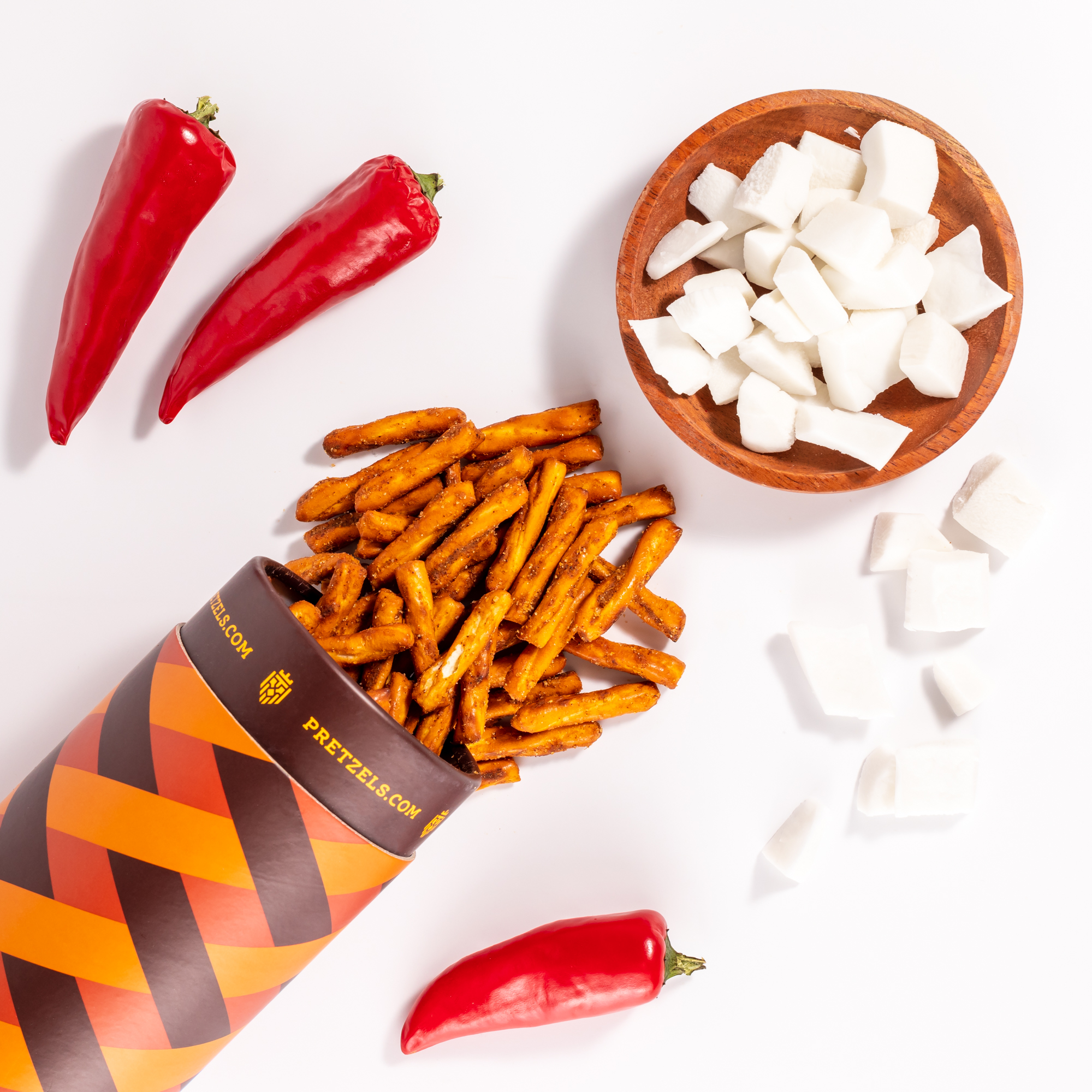 An overhead shot of seasoned pretzels, red peppers, and chunks of coconut on a white background.
