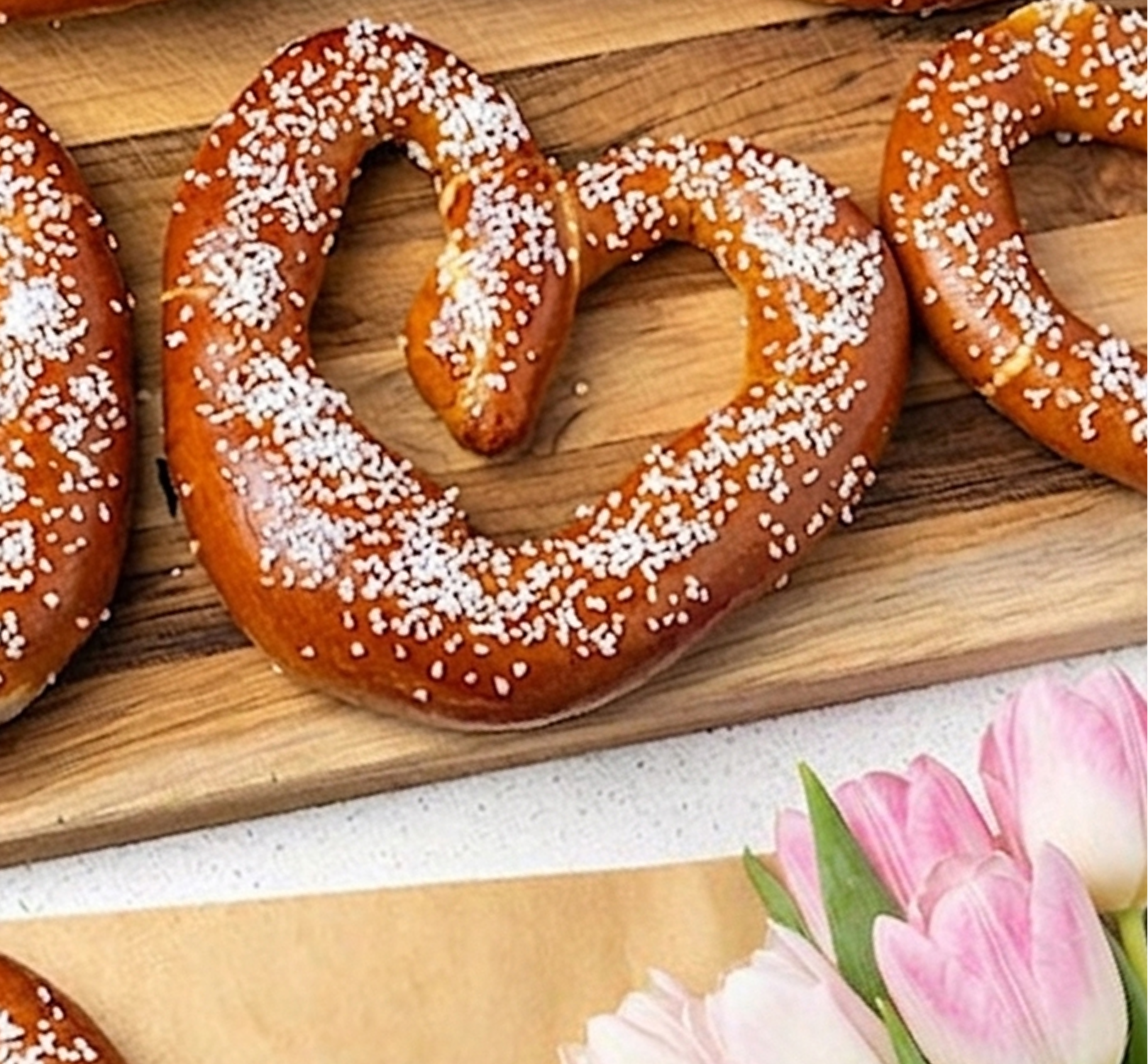 A heart-shaped pretzel with coarse salt on a wooden board next to pink tulips.