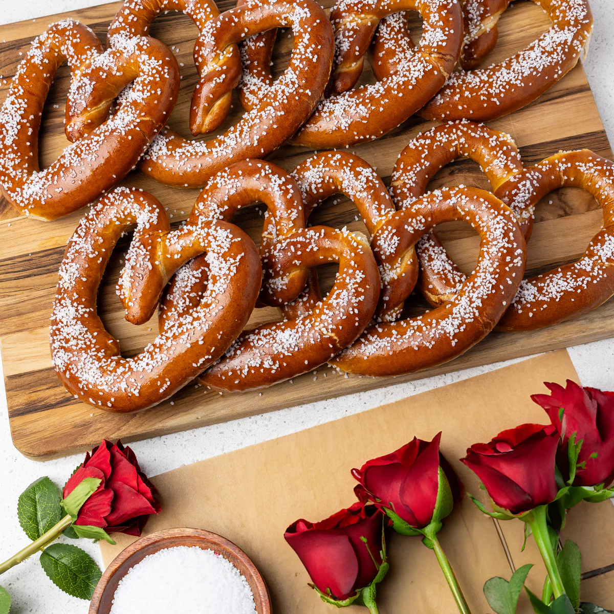 An overhead shot of heart-shaped pretzels on a wooden board next to red roses and a bowl of salt.
