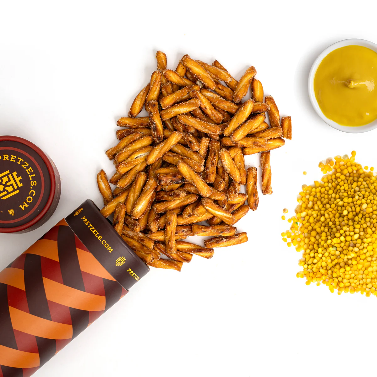 Pretzel sticks spilling from a cylindrical container, with mustard dip and yellow mustard seeds nearby.
