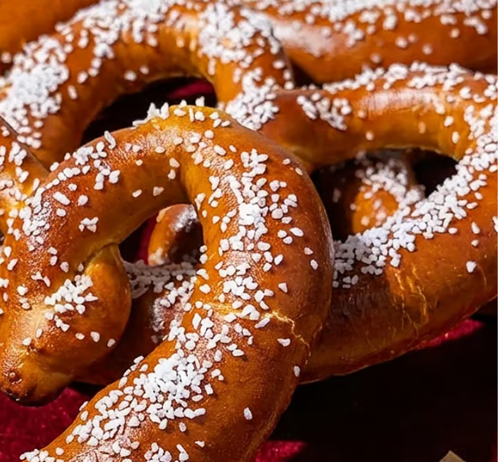 A close-up of several freshly baked soft pretzels generously topped with coarse salt, resting on a red surface.