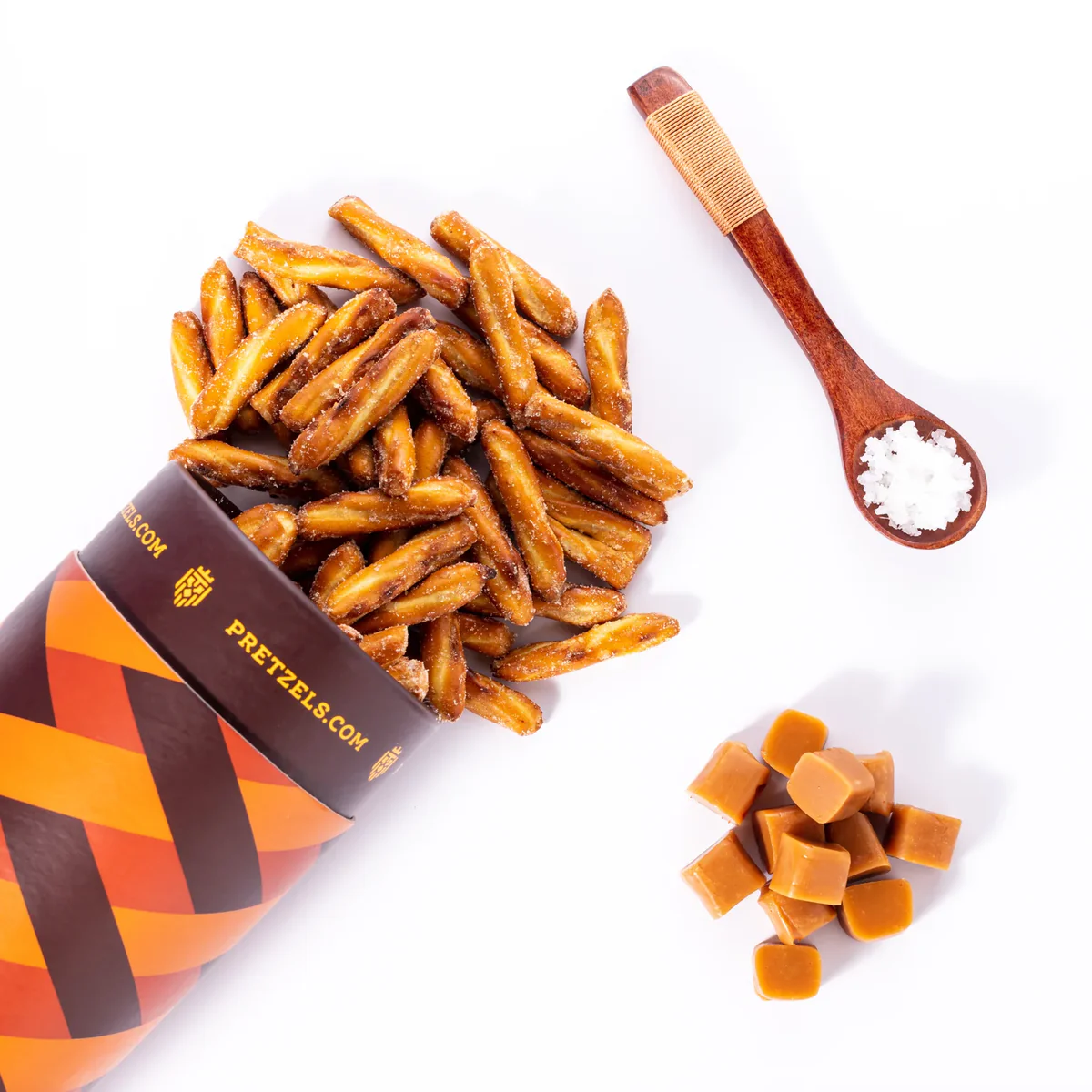 Brown bag spilling pretzels, wooden spoon with salt, and caramel cubes on a white background.